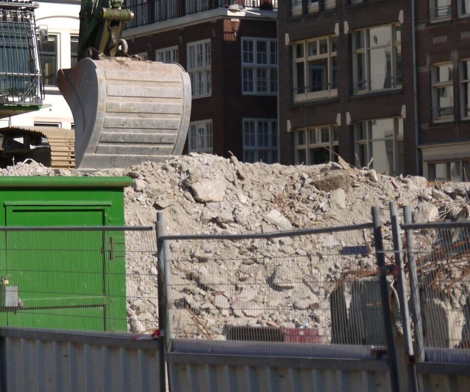 An excavator bucket poised over a pile of broken concrete behind site fencing.