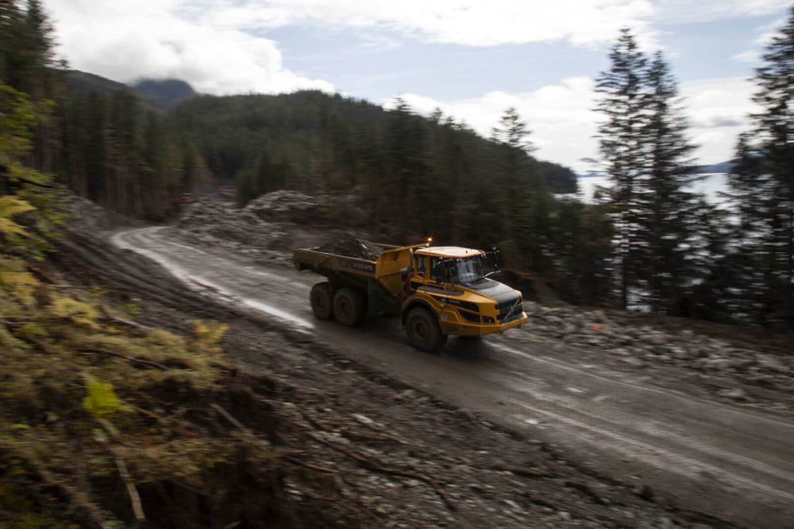 A loaded haul truck on a wet gravel road between stands of conifer.
