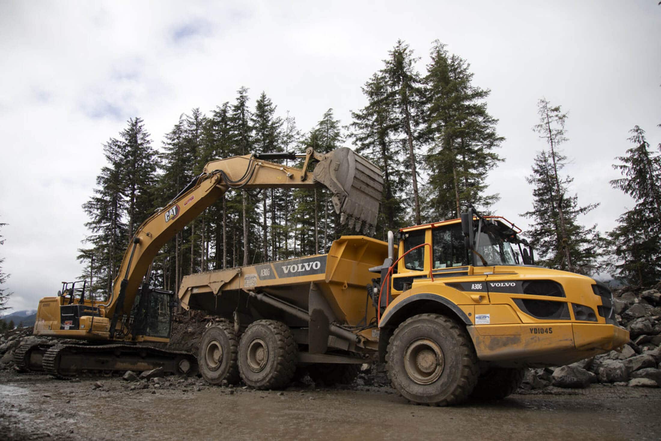 A tracked excavator loading soil into an articulated hauler on a muddy site under an overcast sky.