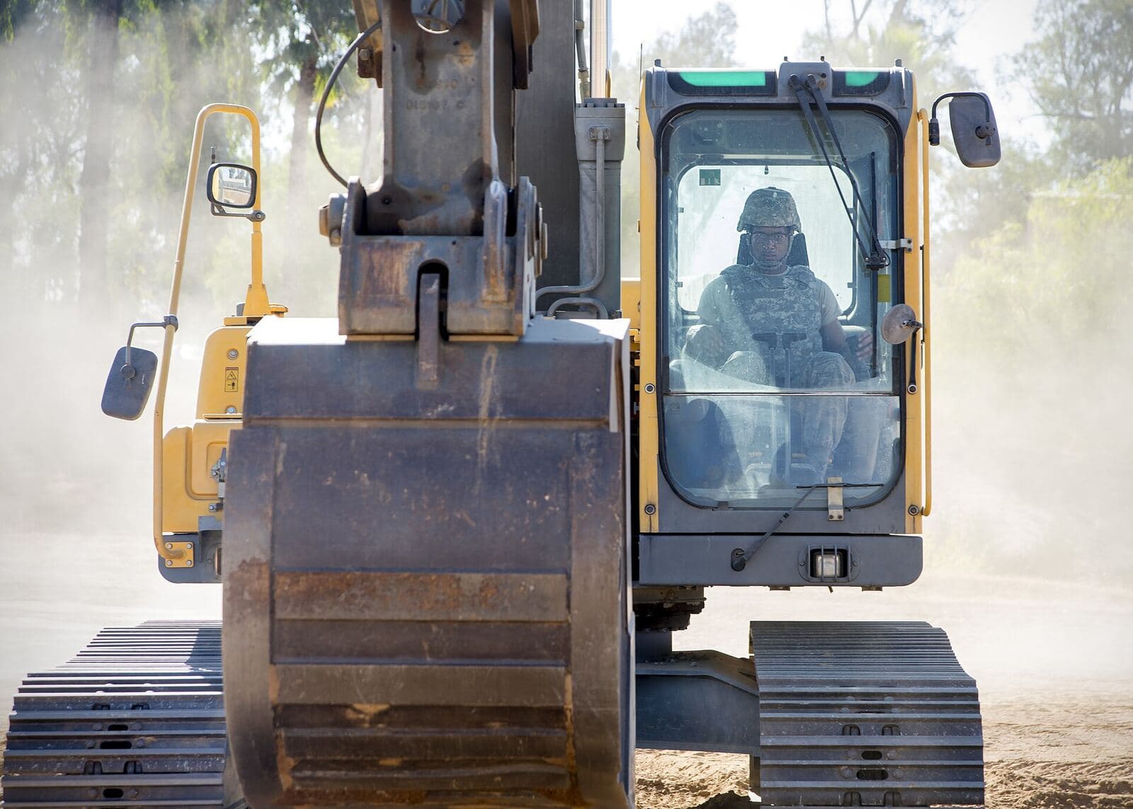 The bucket and cab of a tracked excavator at close range, dust hanging in the air.