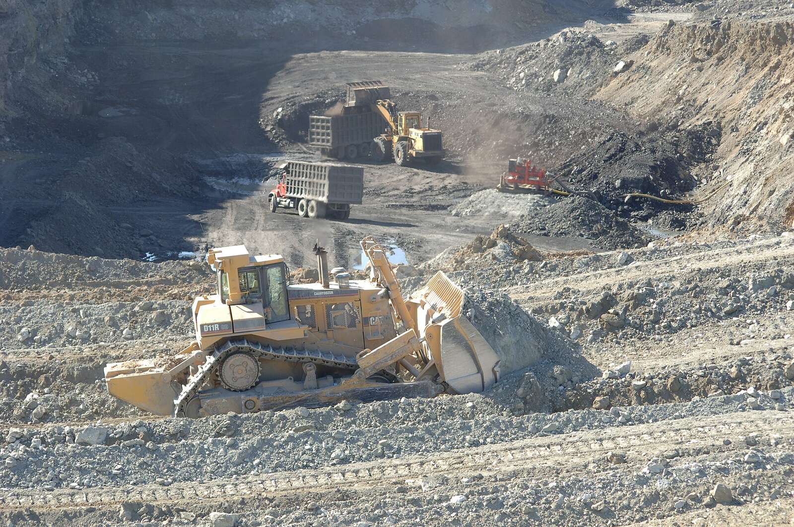 A dozer and haul trucks working a wide bench of broken rock and soil.