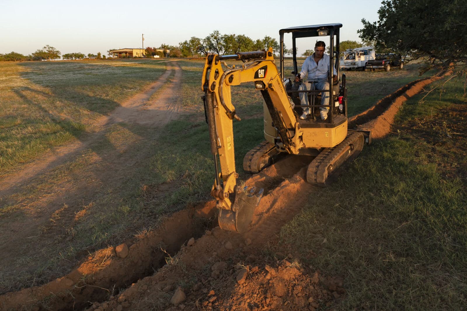 A compact excavator cutting a narrow utility trench across open ground.